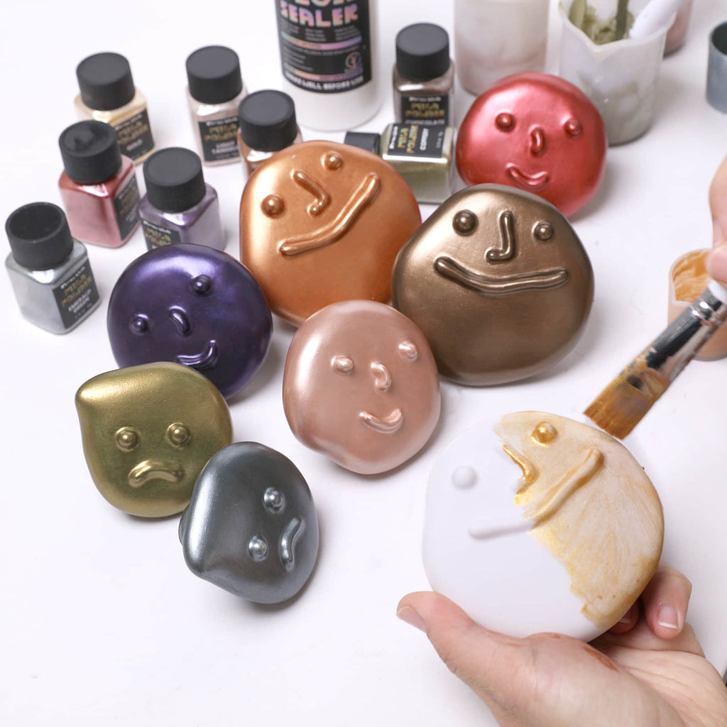 Hand brushing gold onto a white pebble-face wall hook; metallic samples in copper, bronze, purple, and green shown - Boowan Nicole