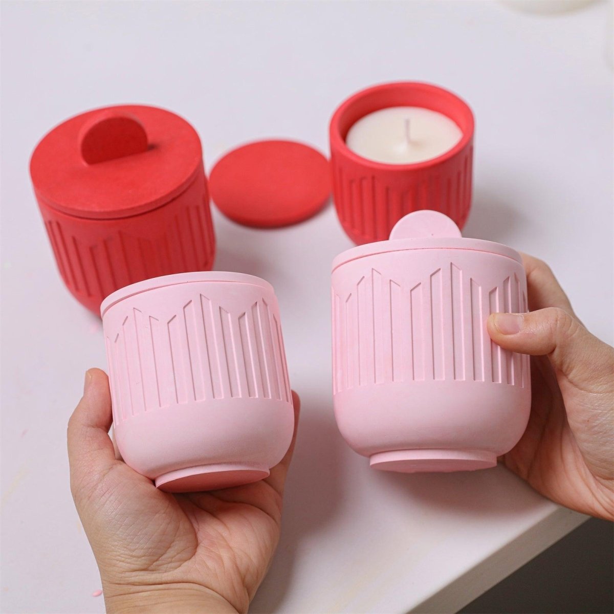 Person holding finished pink candle jars with lids, surrounded by pink jars on table - Boowan Nicole