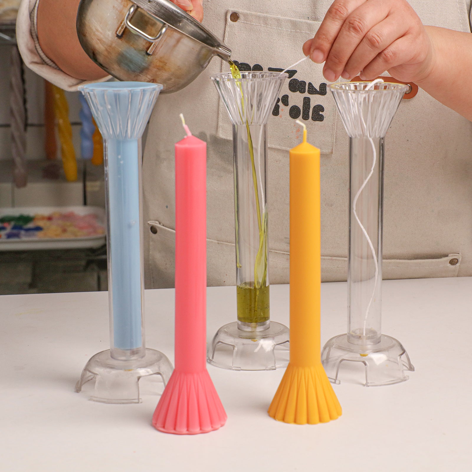 Crafting process: Pouring wax into clear fan-shaped taper candle molds, with finished blue, pink, and yellow candles displayed. -Boowan Nicole