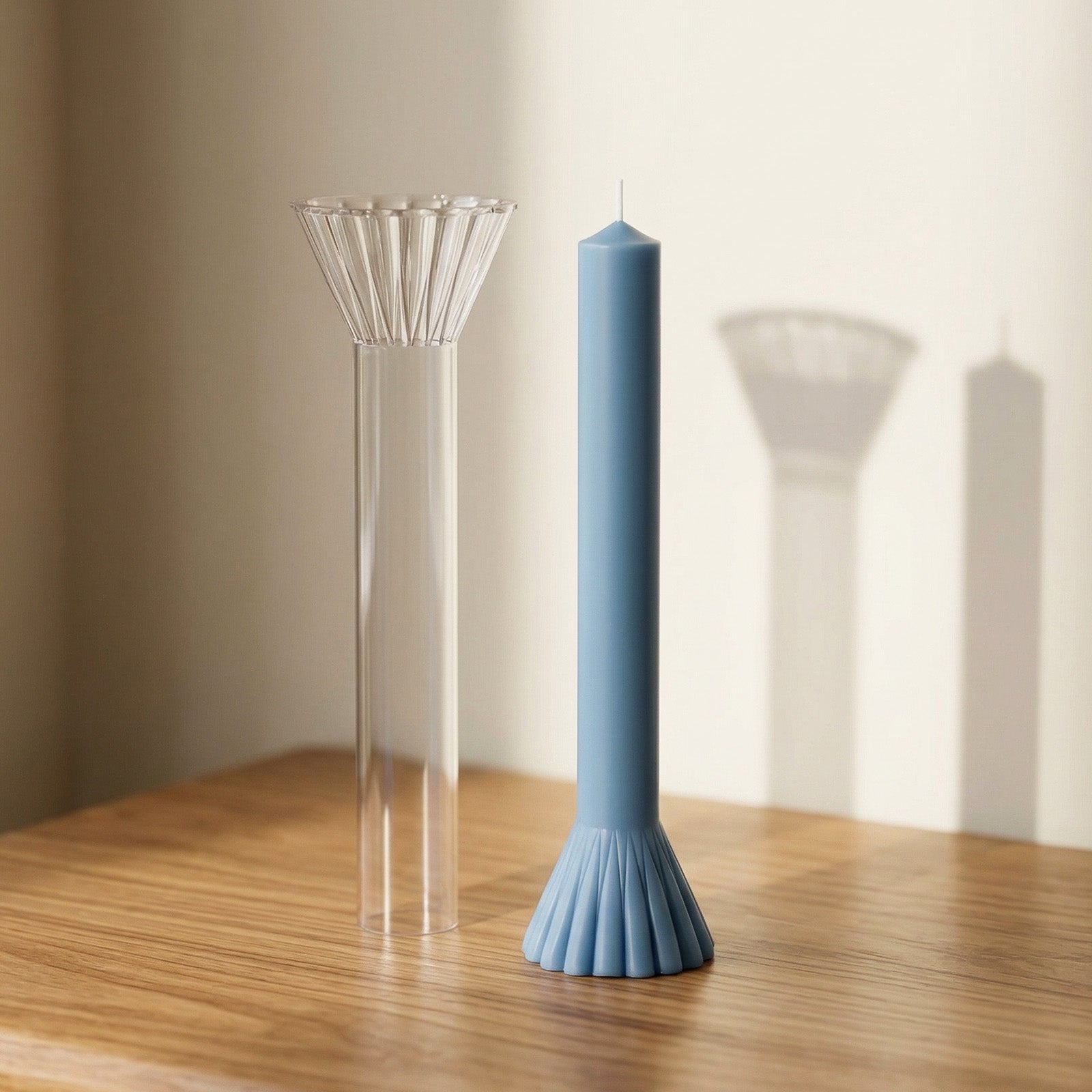 Finished light blue taper candle paired with its clear fan-shaped mold, arranged on a wooden table. -Boowan Nicole