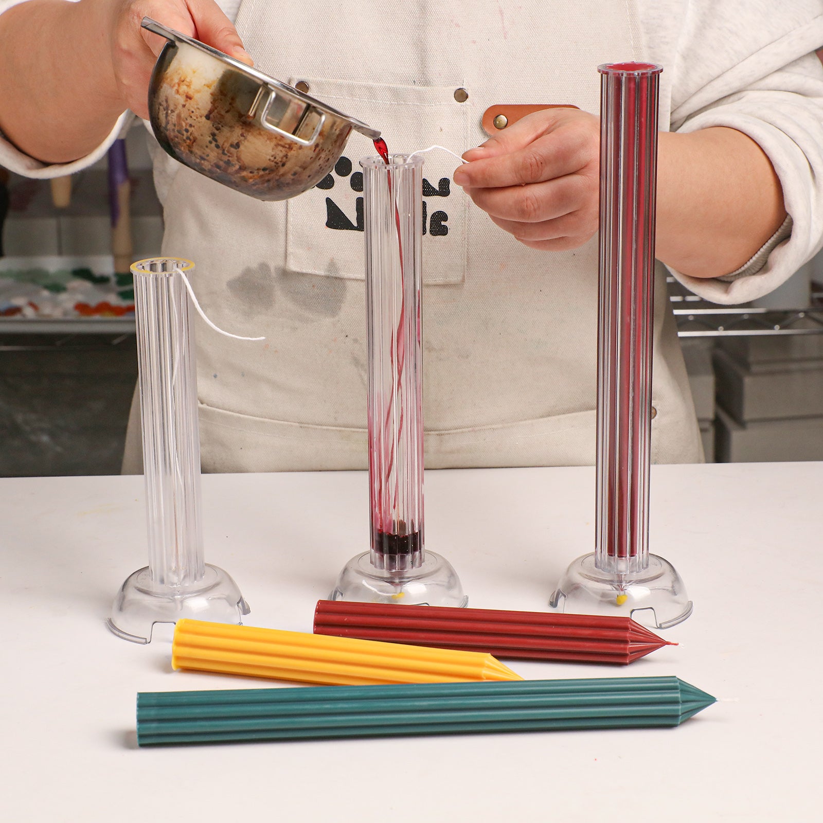 Crafting process: Pouring red wax into a clear ribbed acrylic mold, with finished green, yellow, and red taper candles displayed nearby. -Boowan Nicole