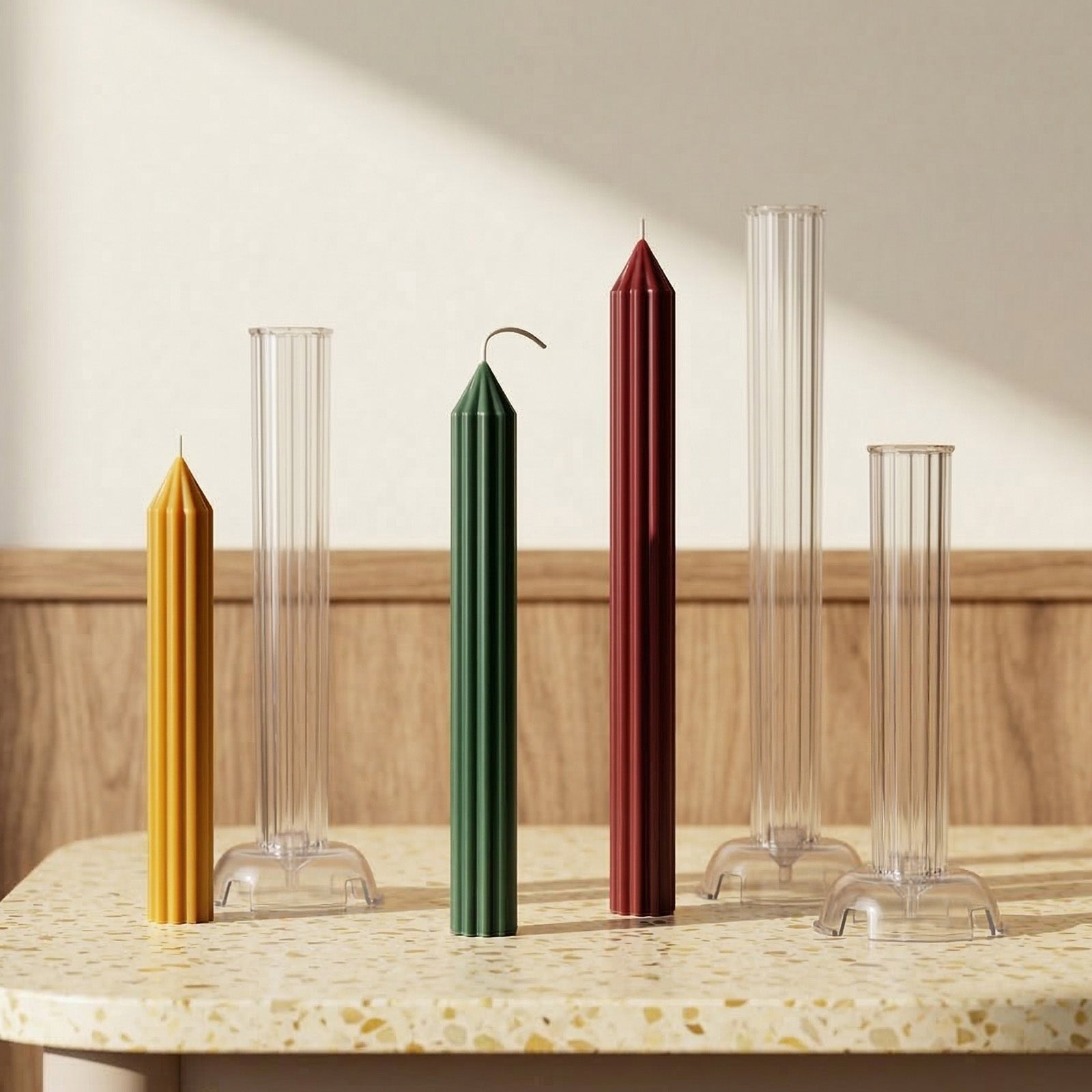 Finished green, yellow, and red ribbed taper candles paired with their clear acrylic molds, displayed on a terrazzo table with soft lighting. -Boowan Nicole
