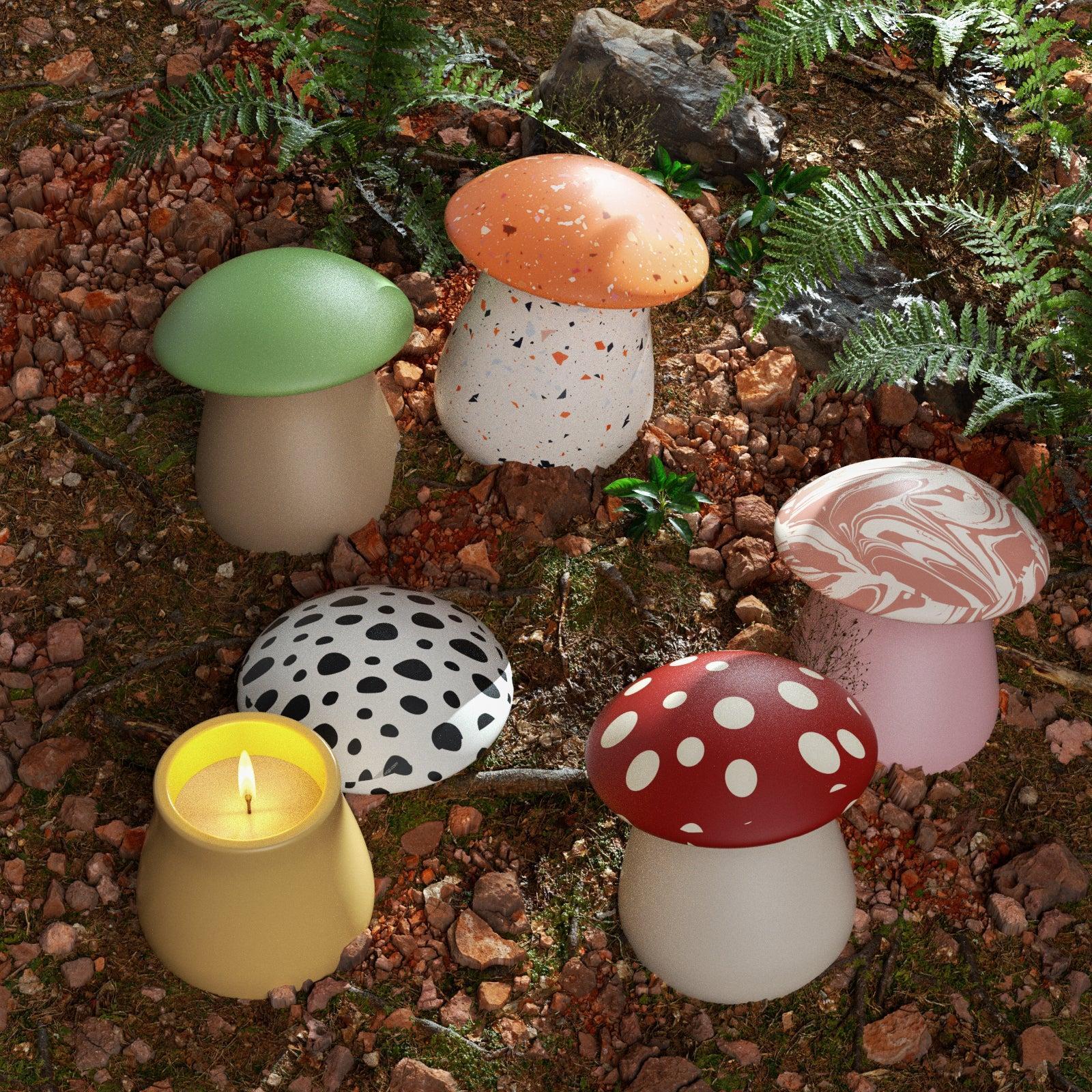 Colorful mushroom-shaped candle jars with patterned lids displayed outdoors on forest ground, one jar lit with a burning candle - Boowan Nicole