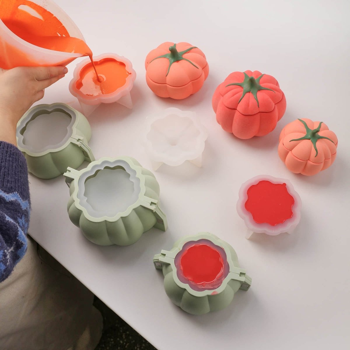 Hand pouring orange wax into tomato-shaped silicone molds with finished tomato candle jars on a white table - Boowan Nicole