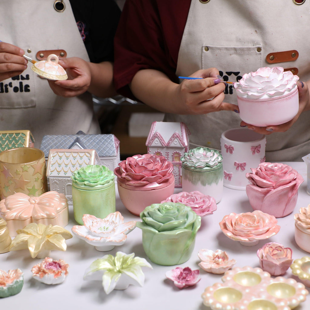 Two crafters painting handmade candle jars shaped like flowers, bows, and houses, decorated with pastel mica powders for DIY home décor - Boowan Nicole