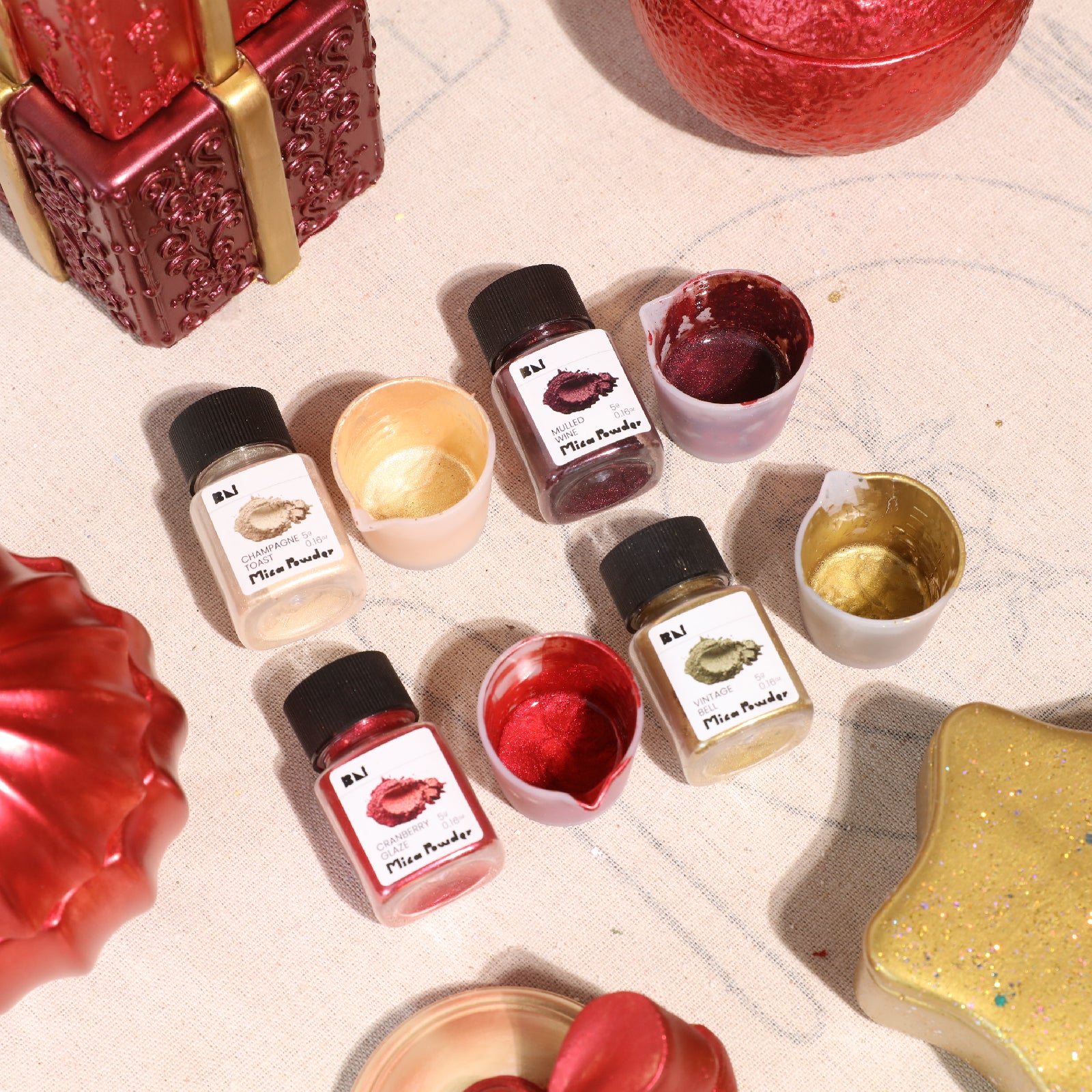 Top-down close-up of 4 Vintage Gala mica powder bottles with mixed metallic paint cups and finished red-gold Christmas crafts - Boowan Nicole