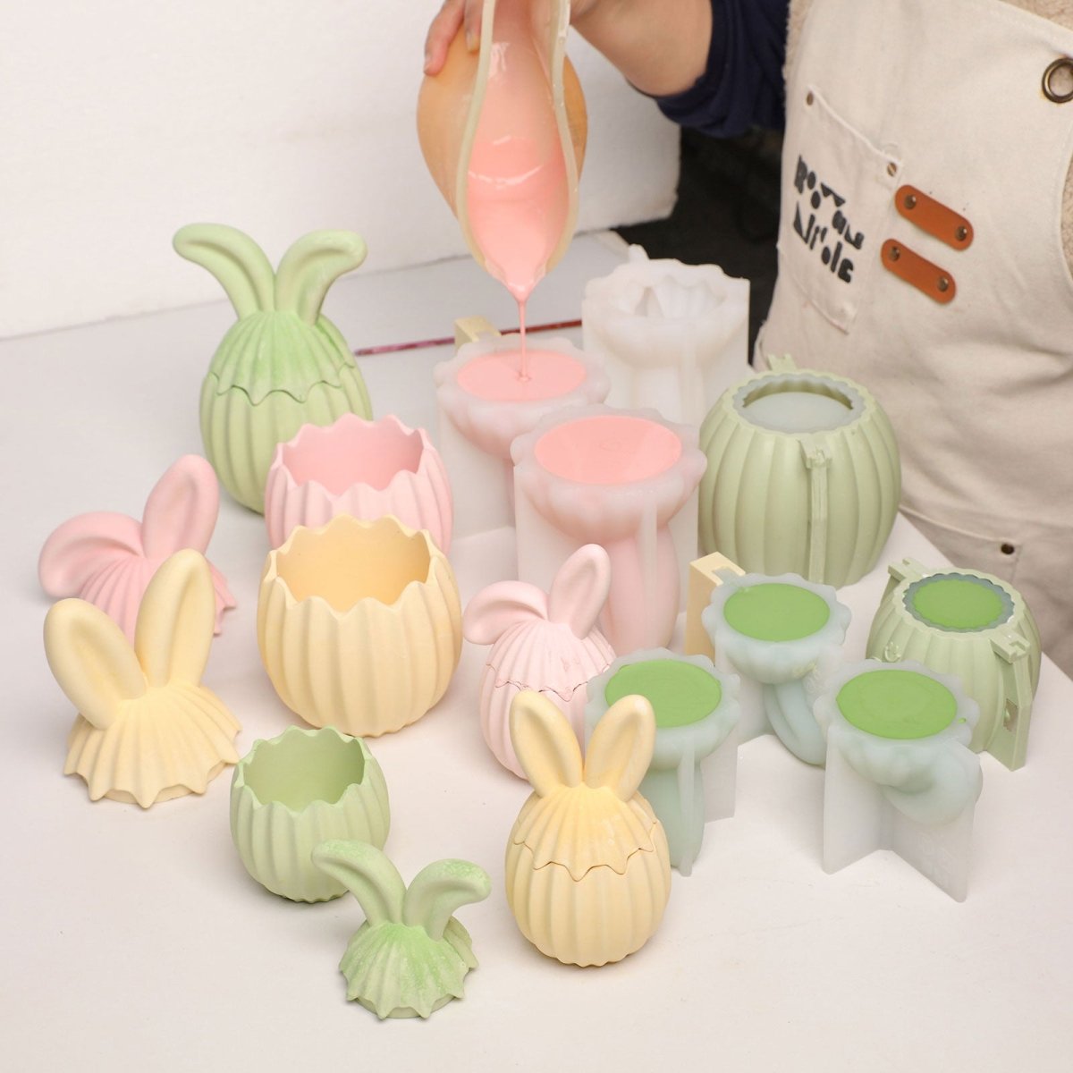 Pouring pink liquid into a silicone mold to create a ribbed bunny ear egg jar, surrounded by finished pastel samples - Boowan Nicole