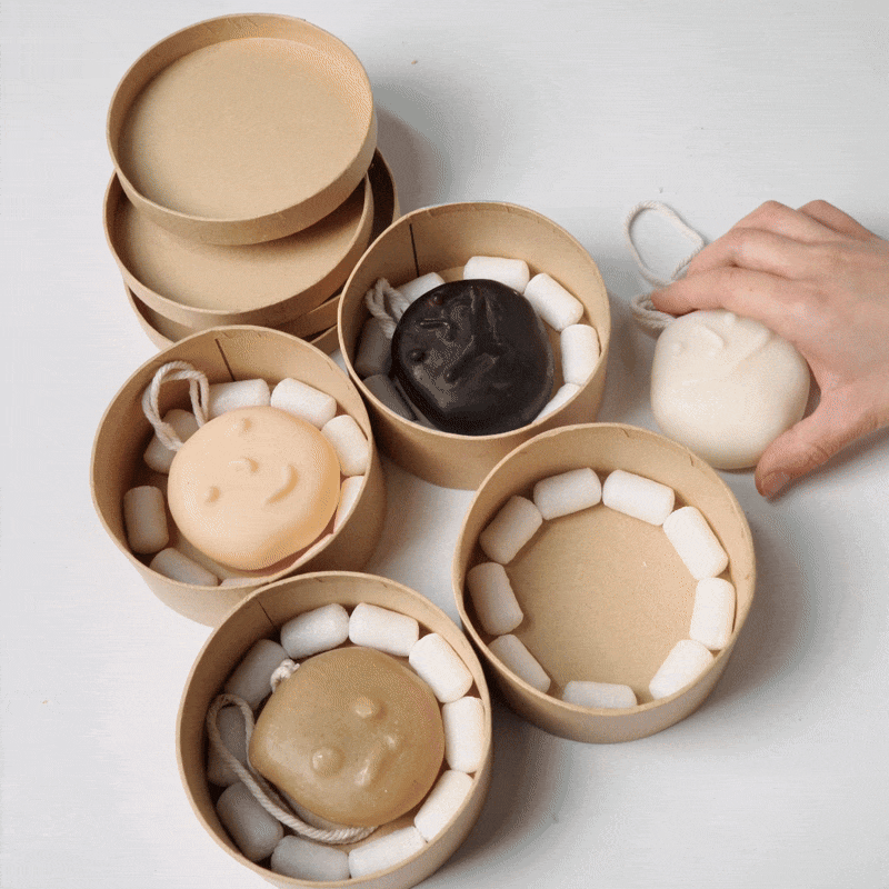 Hand placing a smiley face soap into a kraft packaging box with protective fillers. - Boowan Nicole