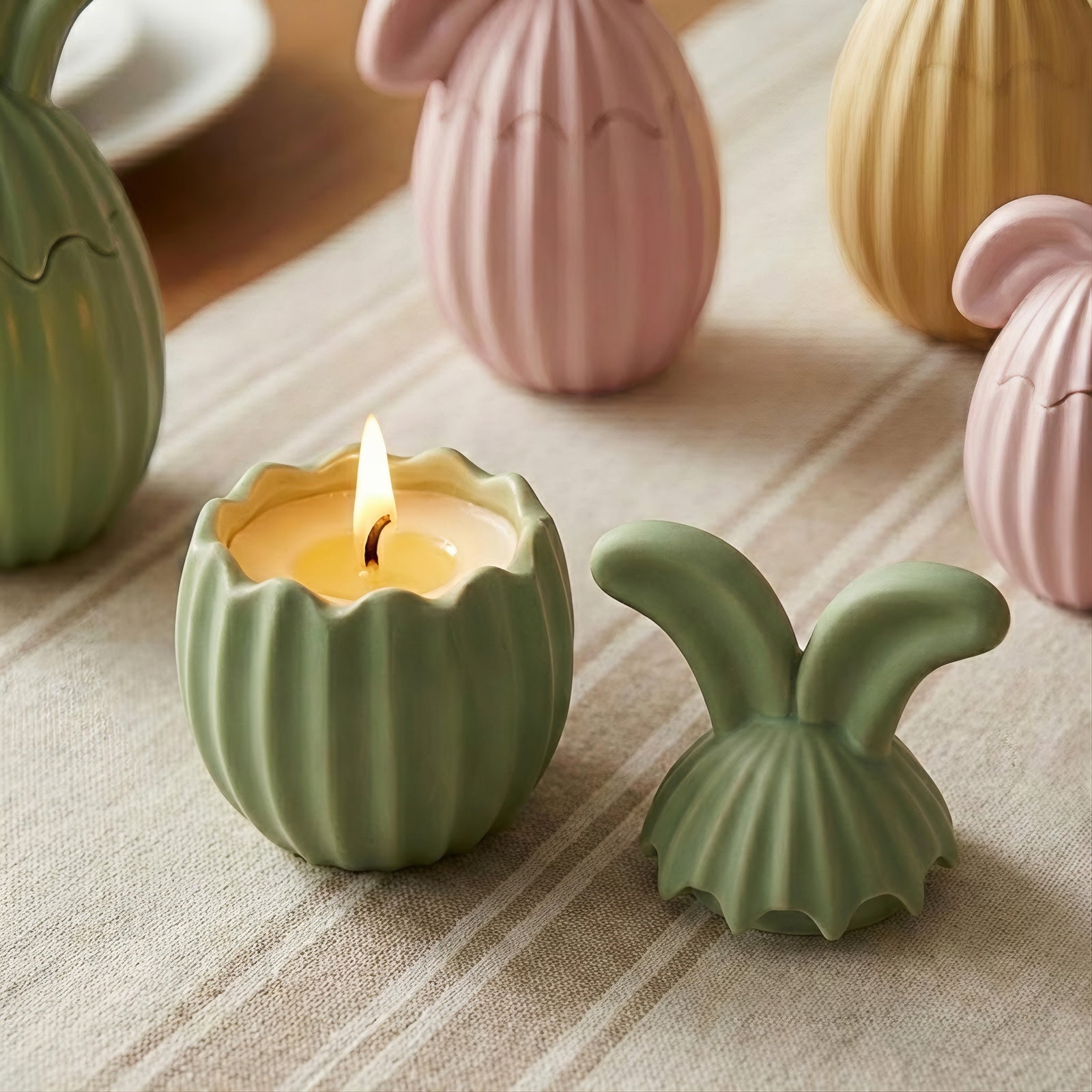 Close-up of an open green bunny ear jar with vertical ribs and a burning candle inside on a beige runner - Boowan Nicole