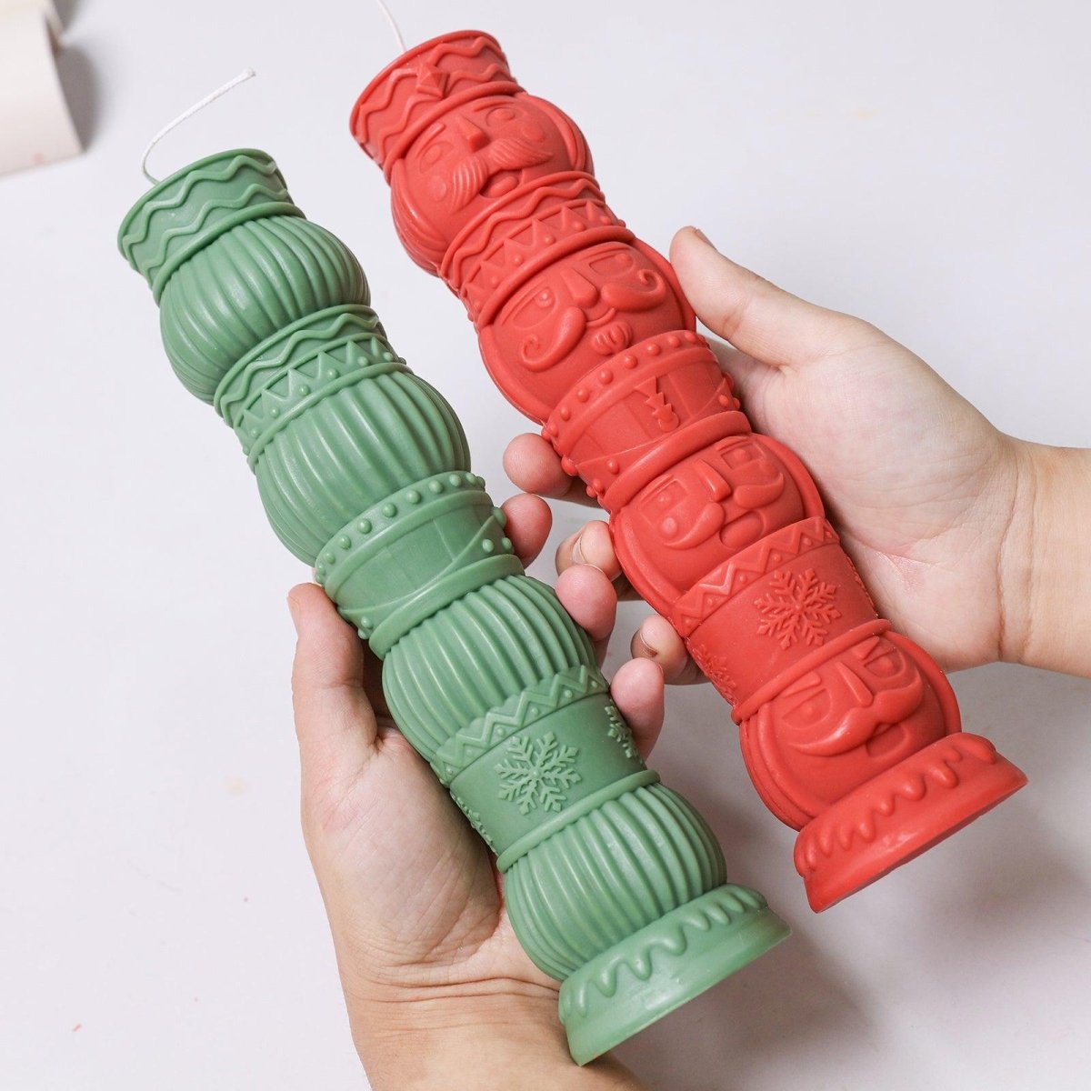 Holding two tall pillar candles with Christmas-themed designs – one in nutcracker totem style, the other with ornament textures, in red and green - Boowan Nicole