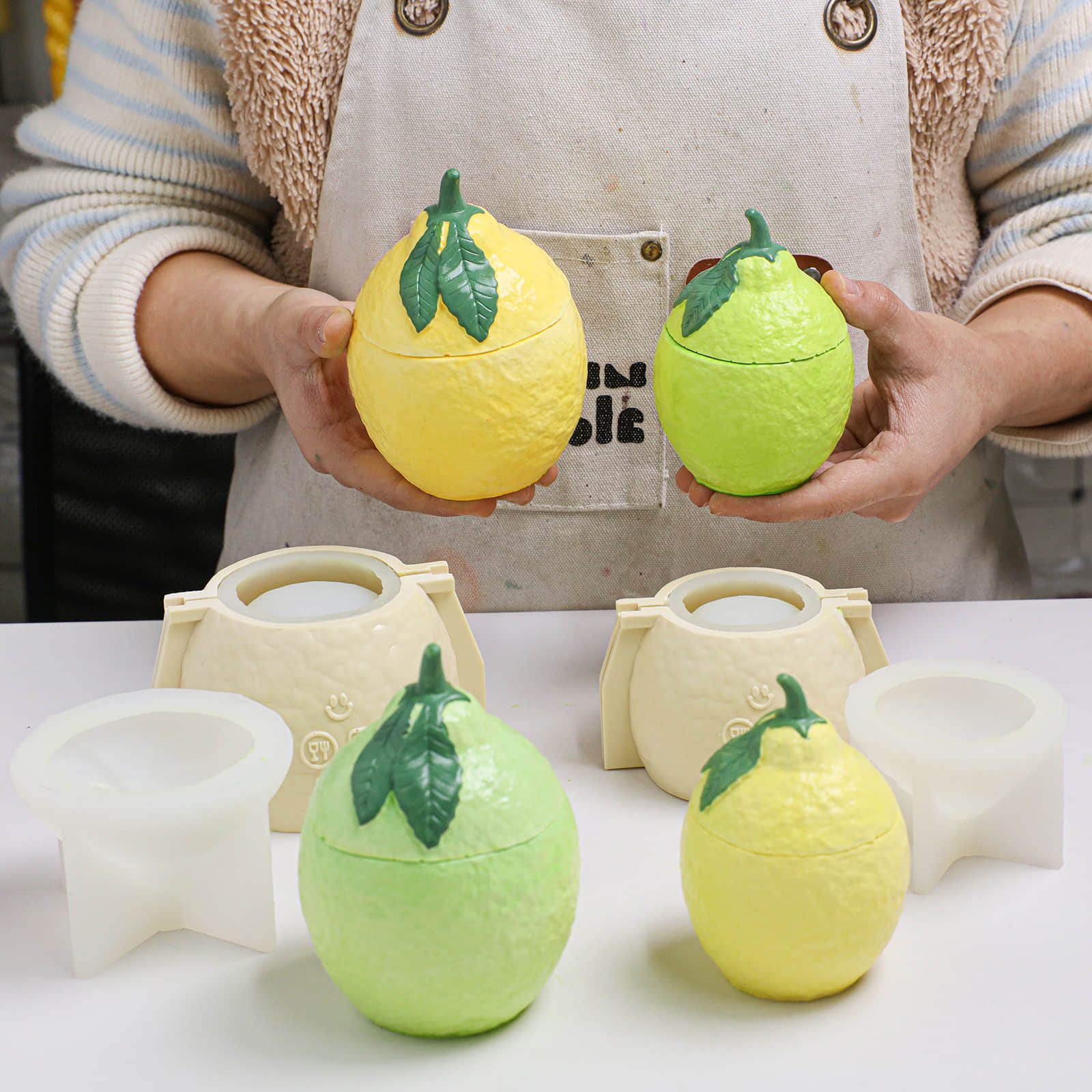 Hands holding yellow and green lemon-shaped candle jars; finished pieces and silicone molds with support shells on the table - Boowan Nicole