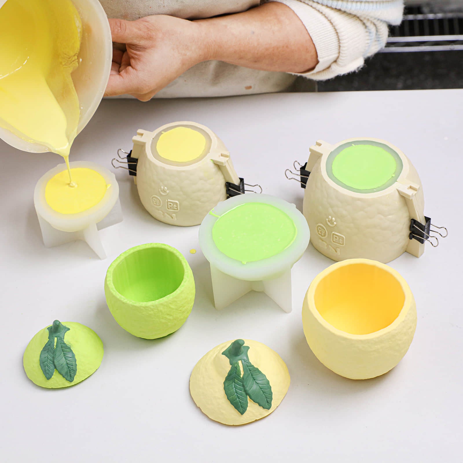 Pouring yellow wax into lemon-shaped candle jar silicone molds with support shells; leaf lids and cured pieces nearby - Boowan Nicole