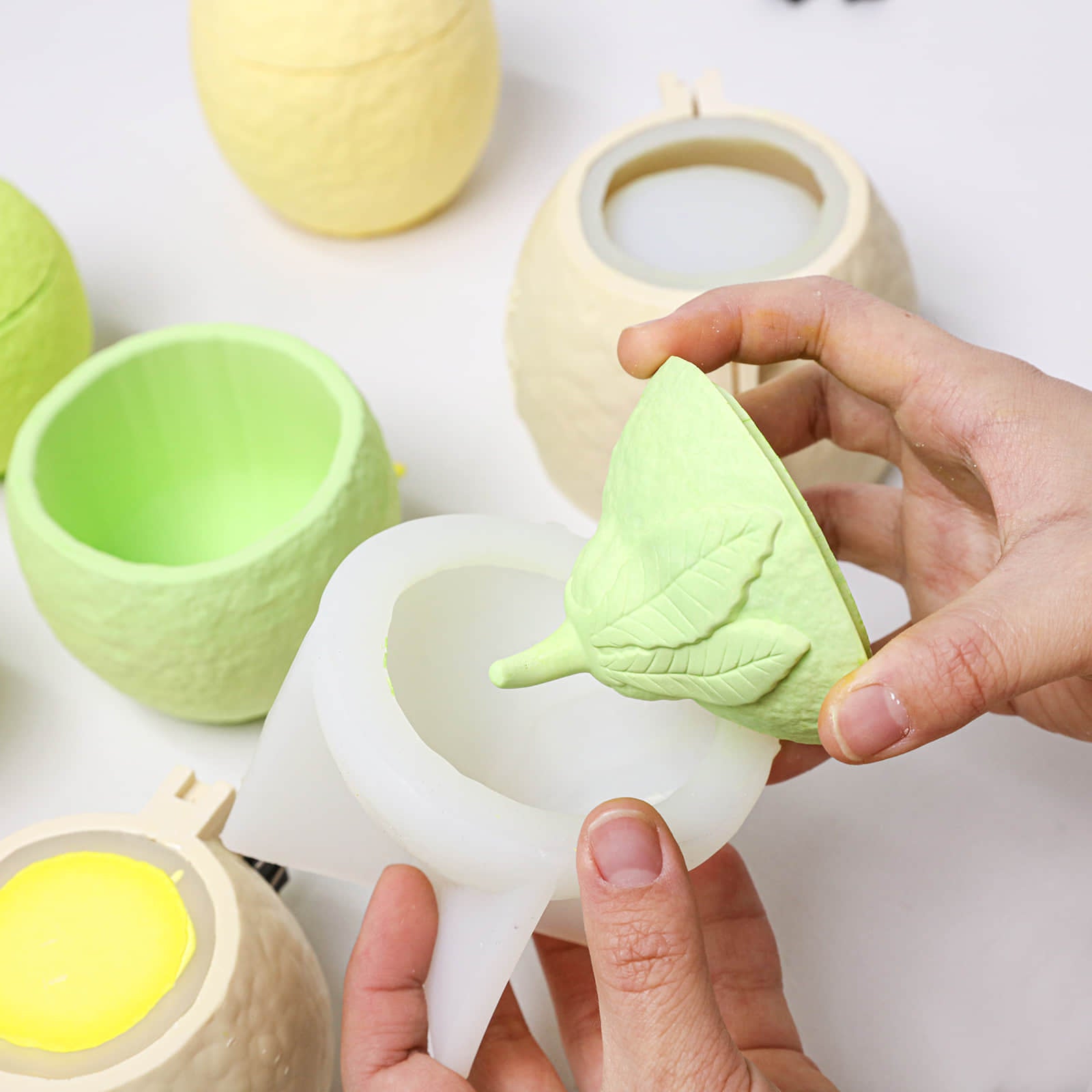 Demolding a lemon-shaped candle jar lid with leaf detail from a silicone mold; jar pieces and support shells nearby - Boowan Nicole