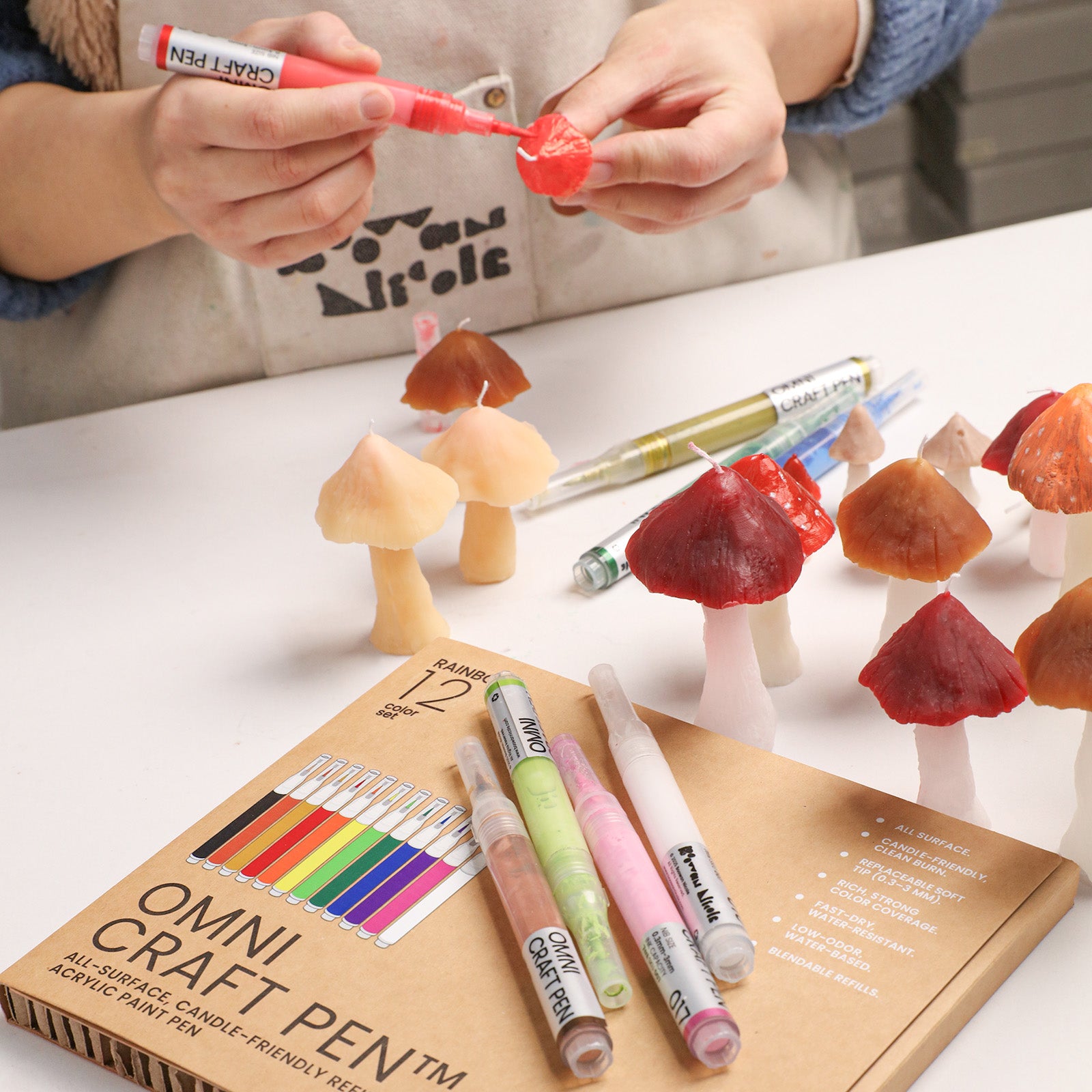 Hand-painting a mushroom-shaped candle using a red OMNI Craft Pen, with a set of acrylic paint pens and various finished candles on the workspace - Boowan Nicole