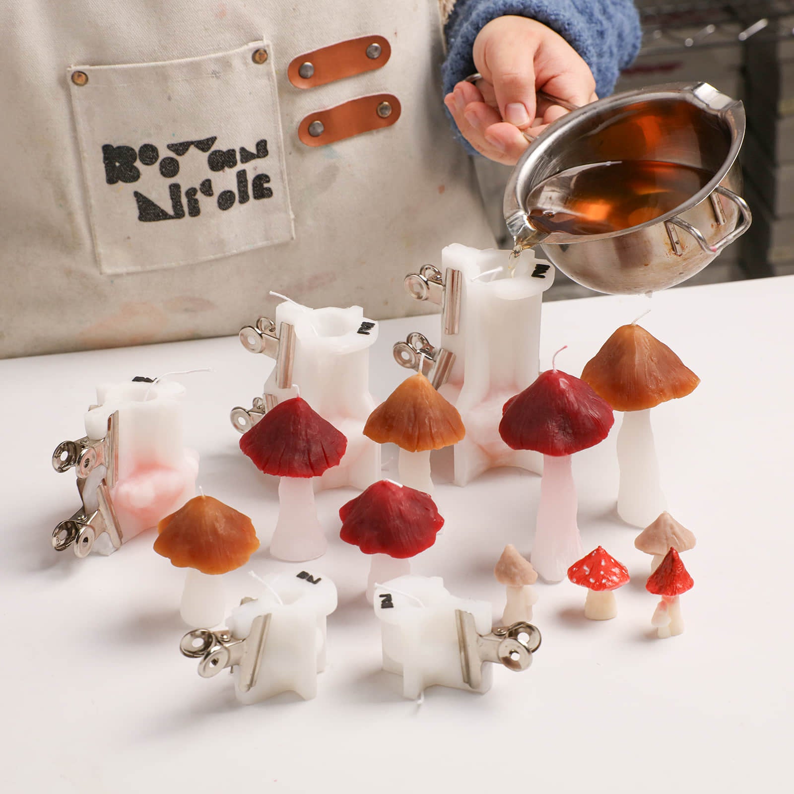 Pouring melted wax into silicone molds, surrounded by finished artisanal mushroom-shaped candles - Boowan Nicole