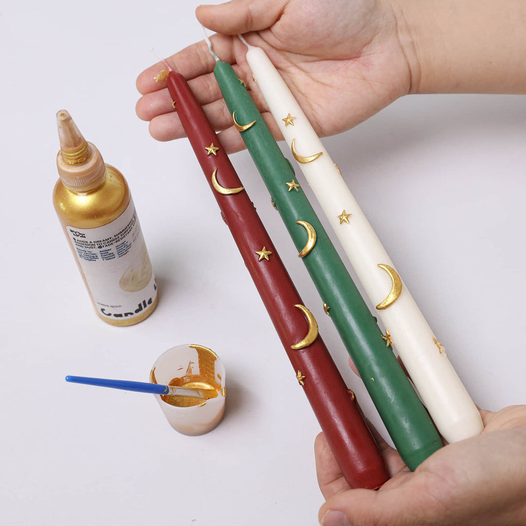 Hands holding red, green, and ivory Midnight Magic taper candles with gold crescent moons and stars; gold varnish and brush on table - Boowan Nicole