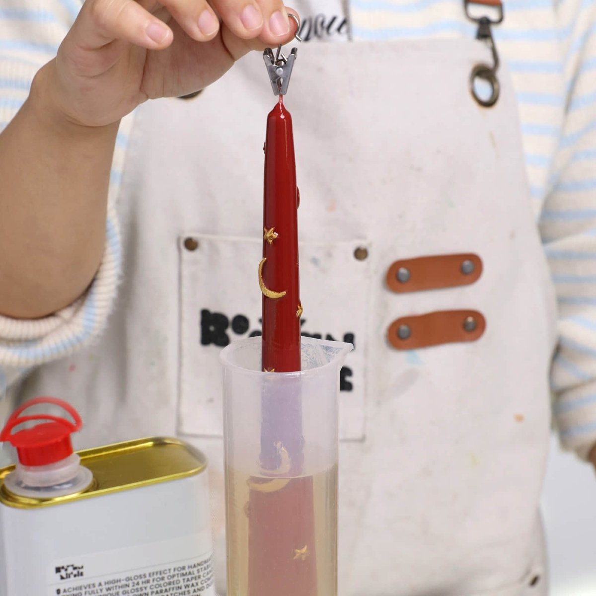 Dipping a red Midnight Magic taper candle—gold crescents and stars—into clear gloss varnish with a clamp - Boowan Nicole