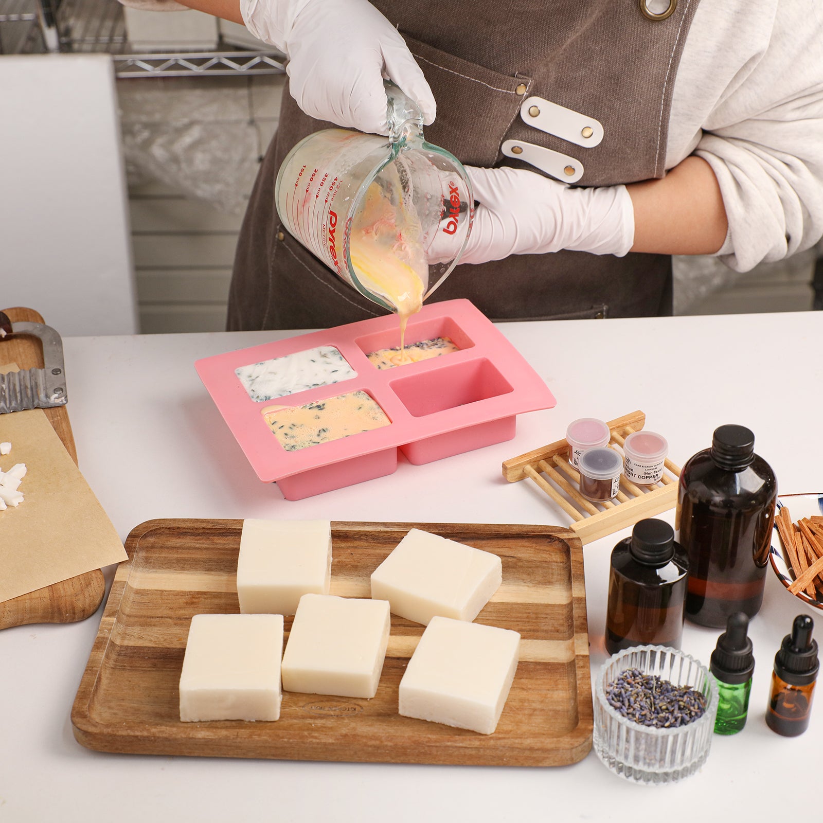 Pouring Milk Cleansing Essential Oil Soap mixture into a mold with finished creamy soap bars - Boowan Nicole