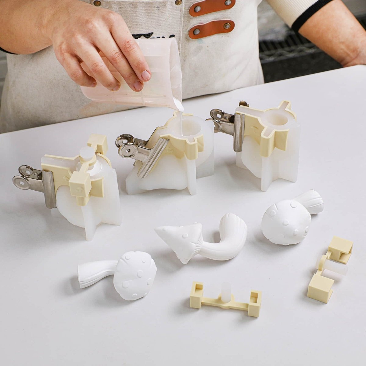 Artisan pouring casting mix into clamped Mushroom Buddies silicone molds, with mushroom hook castings and core inserts on the worktable - Boowan Nicole