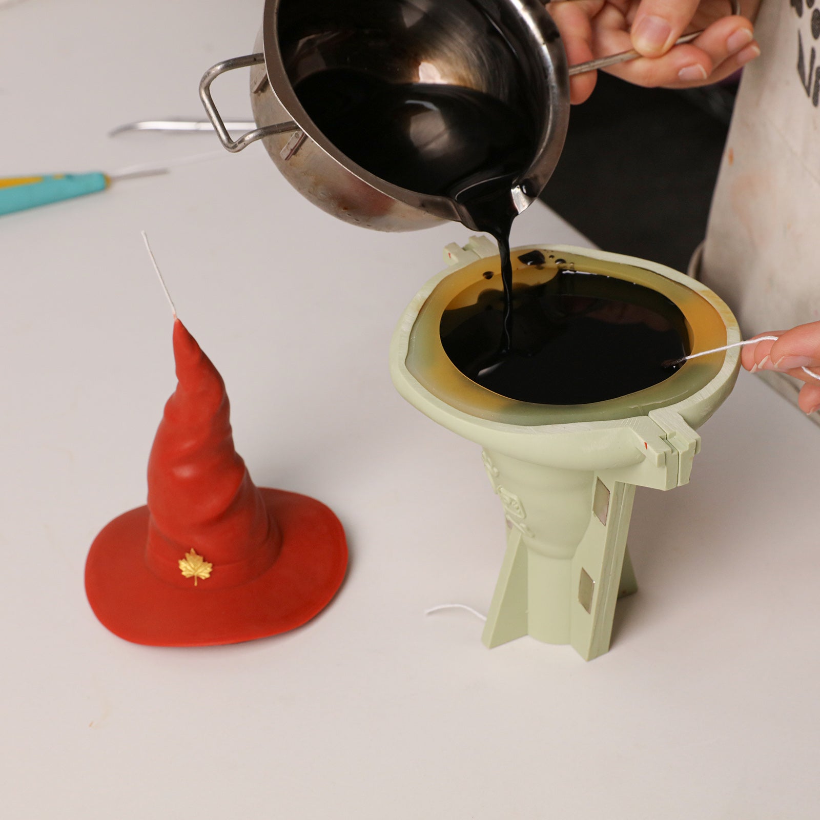 Craft process shot: pouring pigmented wax into mystic wizard hat silicone candle mold beside finished red hat candle blank - Boowan Nicole