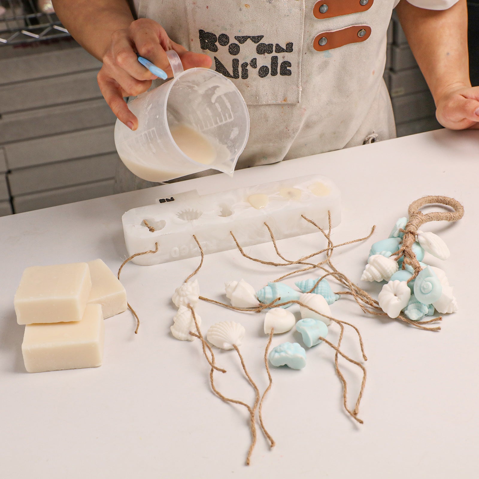 DIY soap making scene, pouring melted soap base into multi-cavity seashell silicone mold with finished shell soap strands - Boowan Nicole