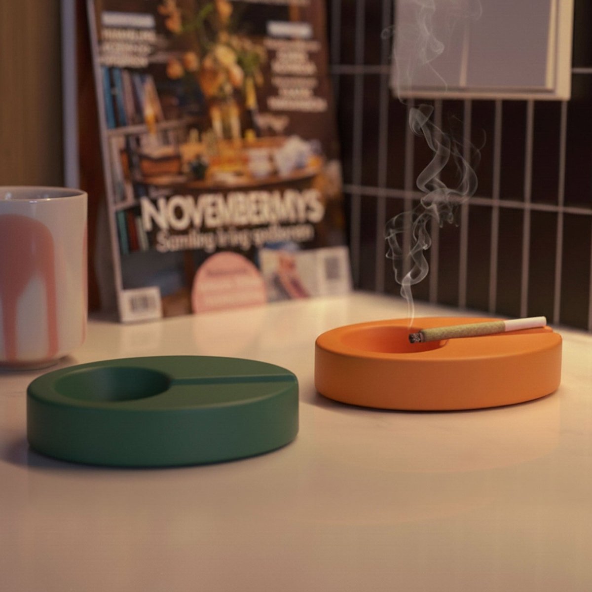 Green and orange Orbit oval ashtrays with bowl and slim groove; one holds a smoking cigarette on a countertop - Boowan Nicole