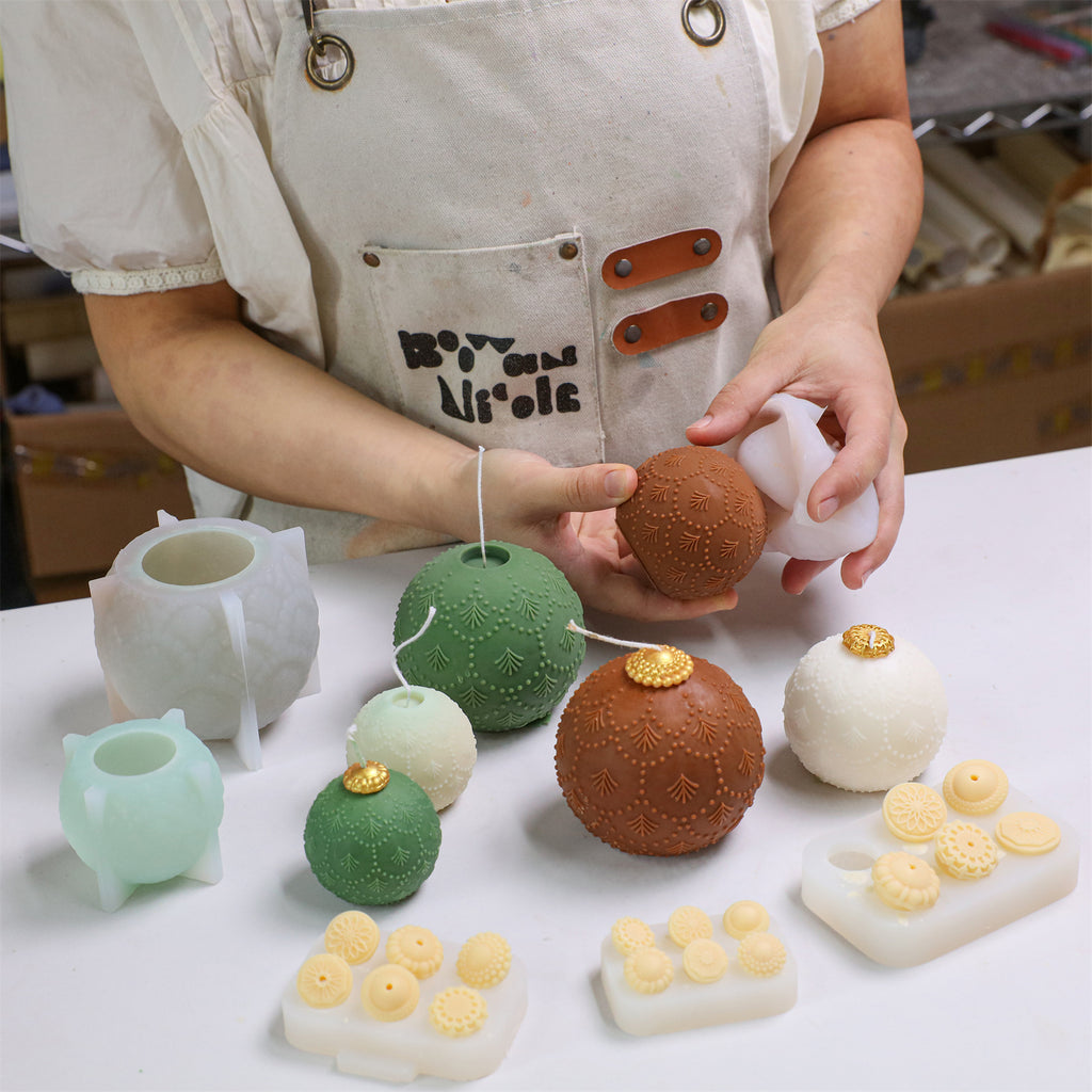 Hands demolding a fir-embossed bauble candle; green, cocoa, and ivory candles with gold caps and topper-cap molds on the worktable - Boowan Nicole