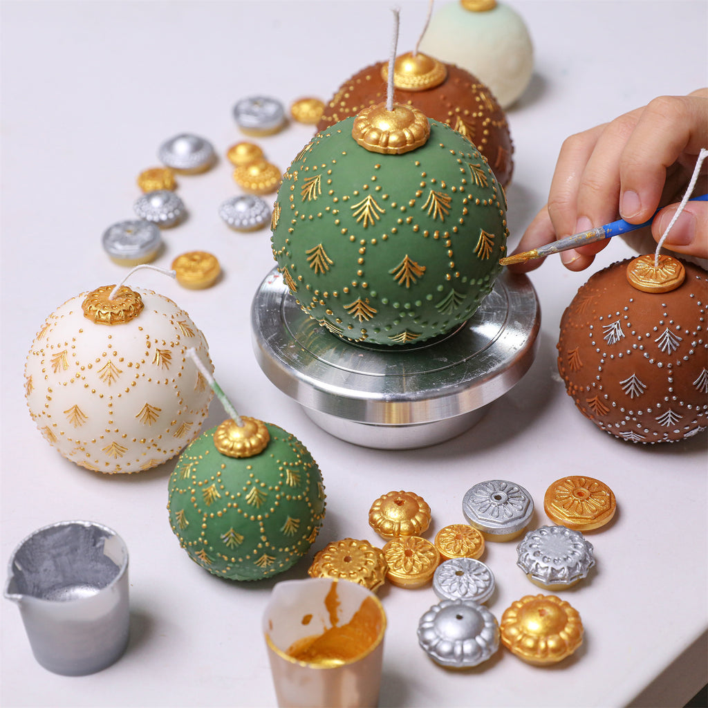Hand painting gold details on a green fir-embossed bauble candle—ivory and cocoa candles and gold/silver topper caps on the worktable - Boowan Nicole