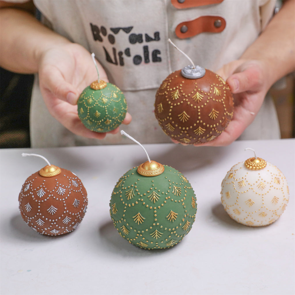 Hands displaying fir-embossed bauble candles—green, cocoa, and ivory—with gold/silver caps; additional finished candles on the table - Boowan Nicole