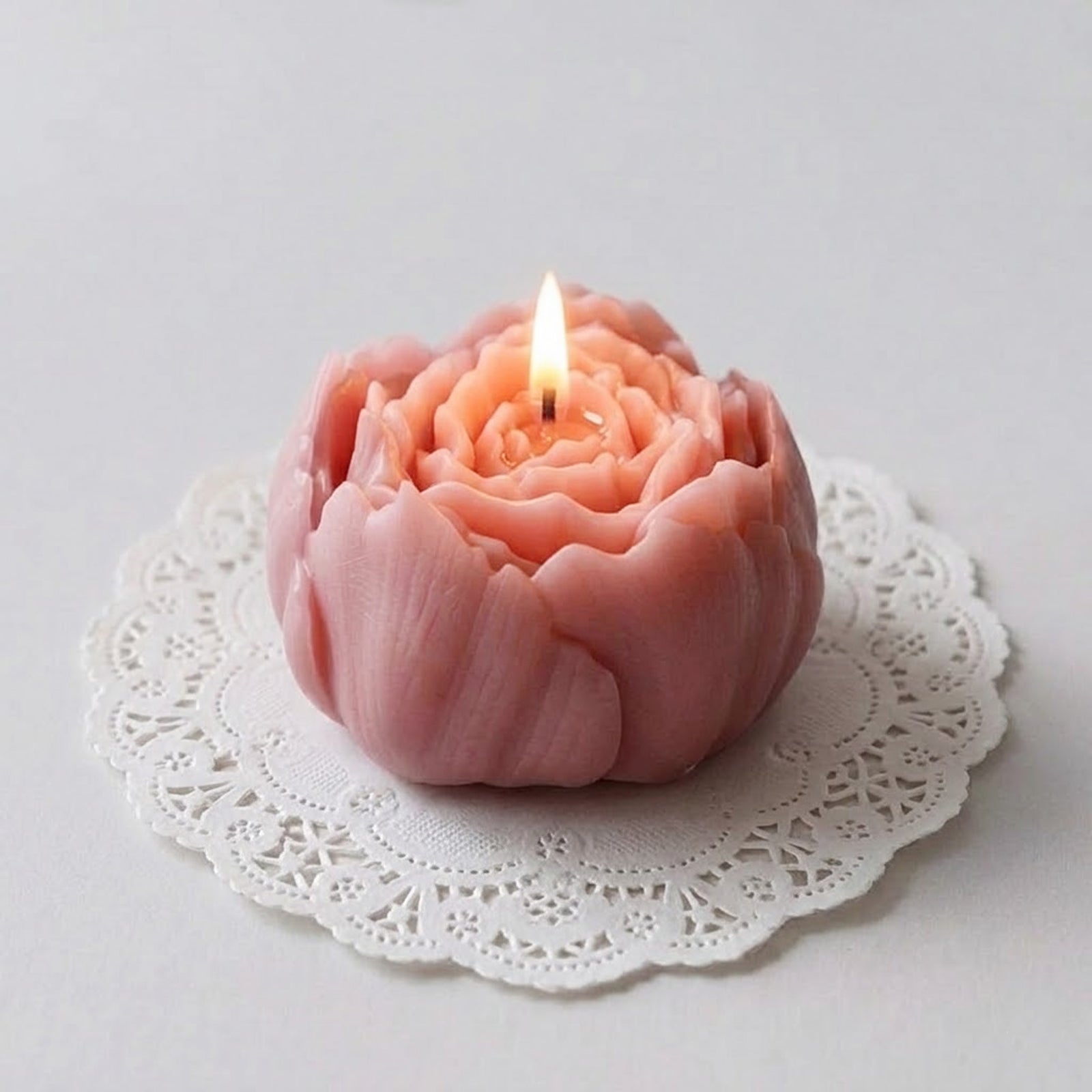 A lit pink peony candle resting on a white lace doily, casting a warm, soft glow. -Boowan Nicole