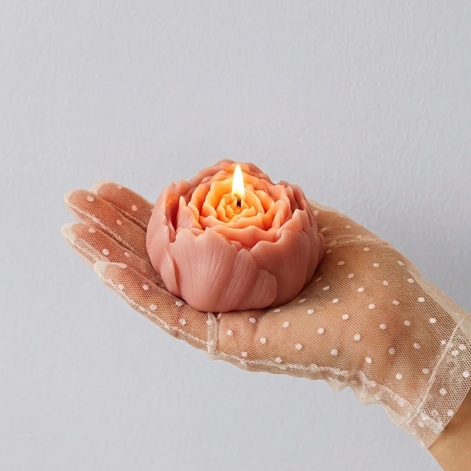 A hand in a polka-dot tulle glove holding a lit pink peony candle, showcasing its elegant floral form. -Boowan Nicole
