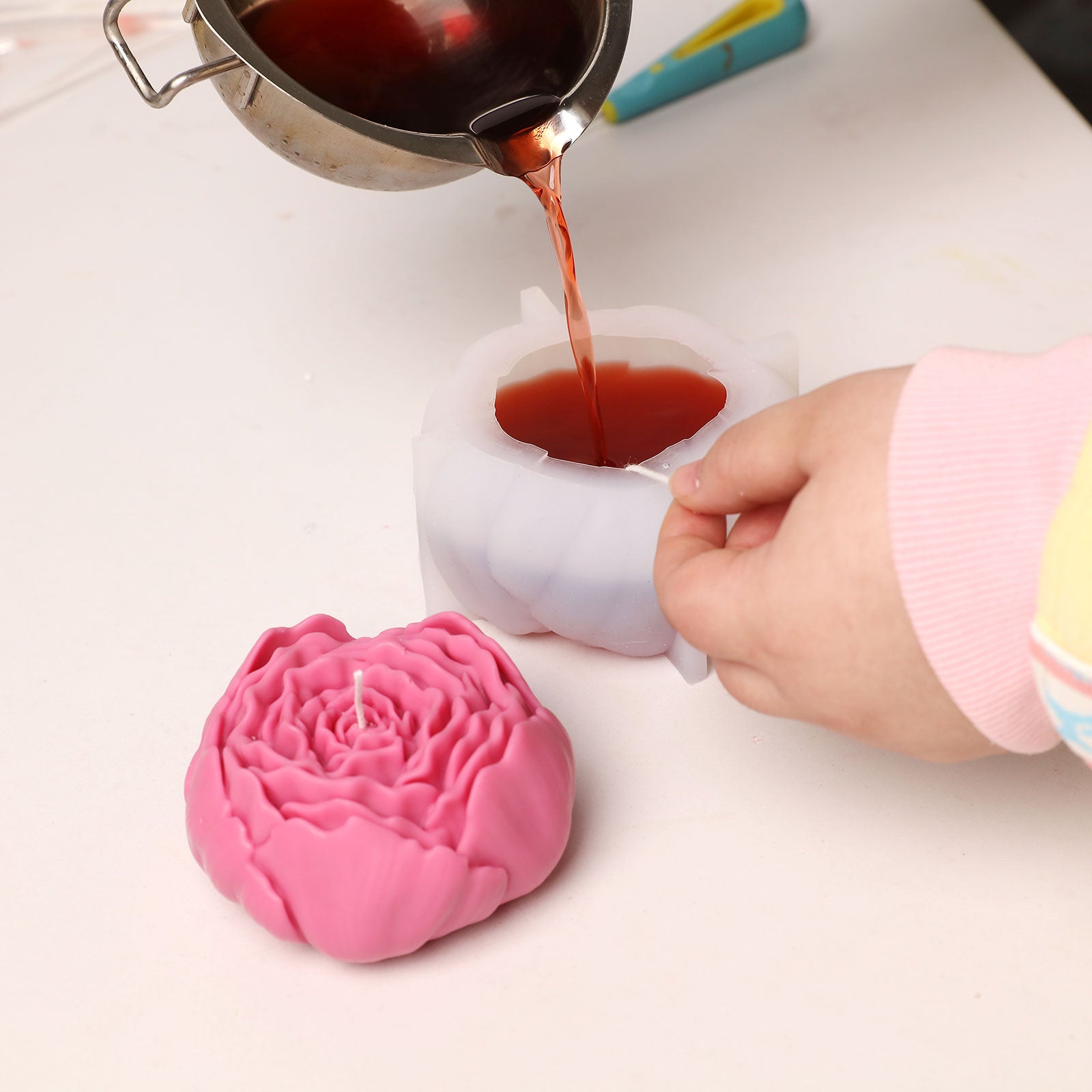 Pouring red wax into a peony-shaped silicone mold, with a finished pink peony candle placed beside it. -Boowan Nicole