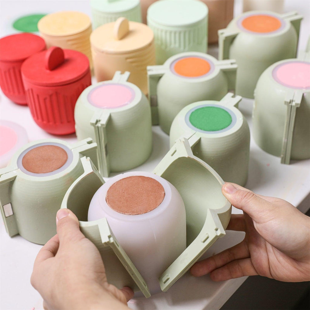 Hands opening a reinforced support shell to release a candle jar from a silicone mold, surrounded by colorful molds - Boowan Nicole
