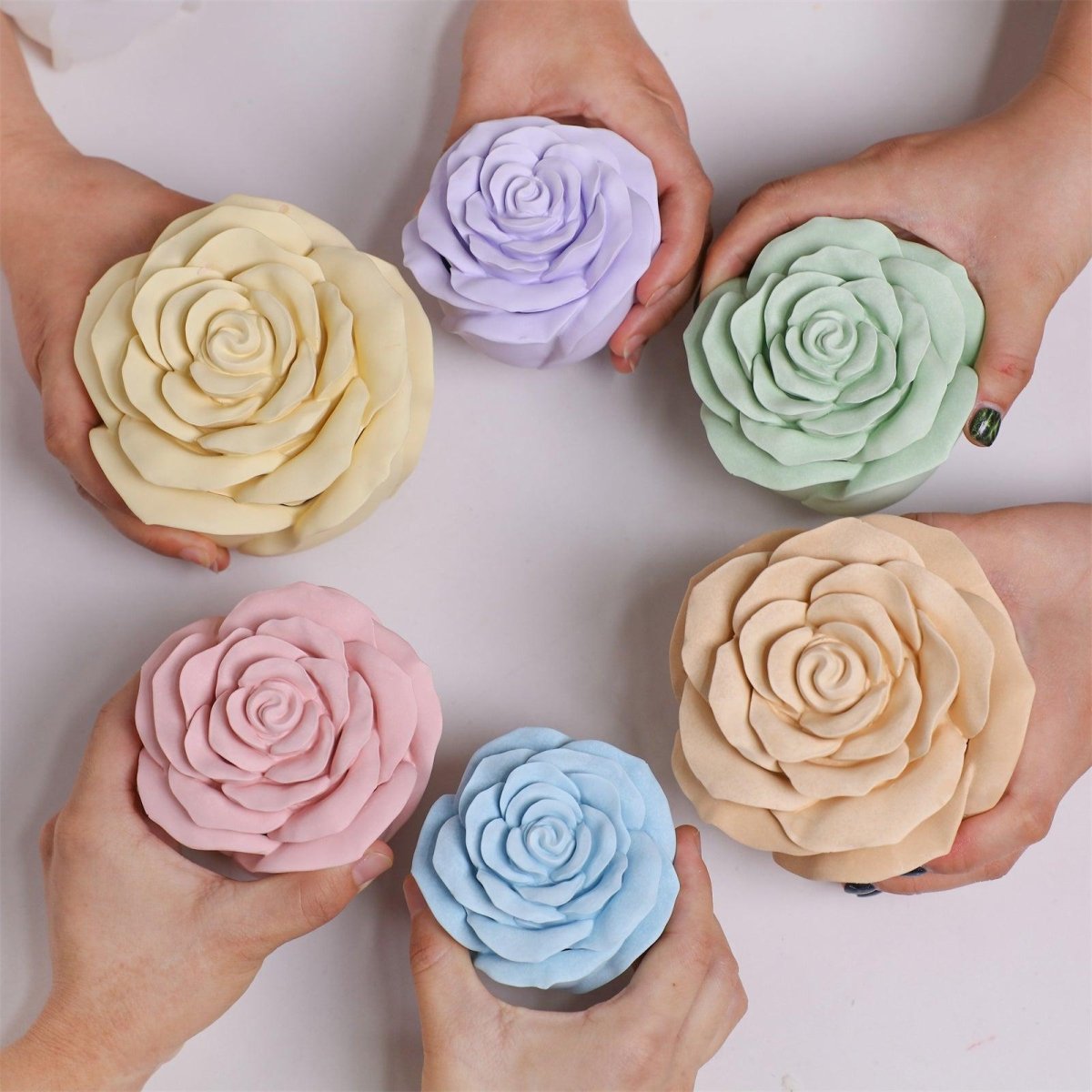 Six hands holding rose-shaped candle jars in various pastel colors: yellow, purple, green, pink, blue, and beige - Boowan Nicole