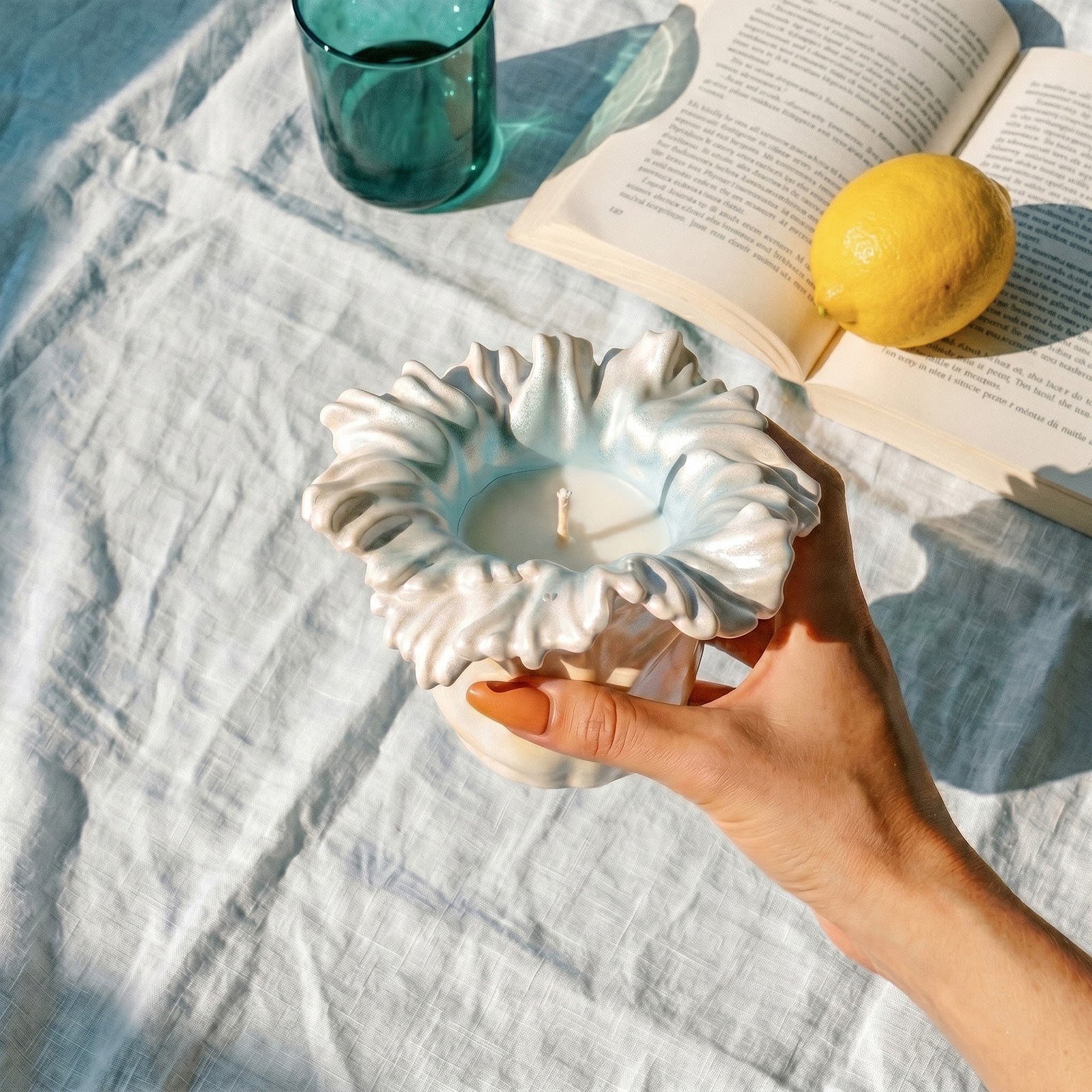 Hand holding iridescent Sea Anemone Bloom Candle Jar with candle, styled with book, lemon and glass - Boowan Nicole