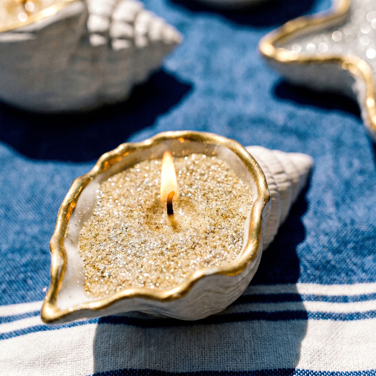 Close-up of Lit Gold Rim Conch Shaped Glitter Sand Wax Candle on Blue White Striped Linen - Boowan Nicole