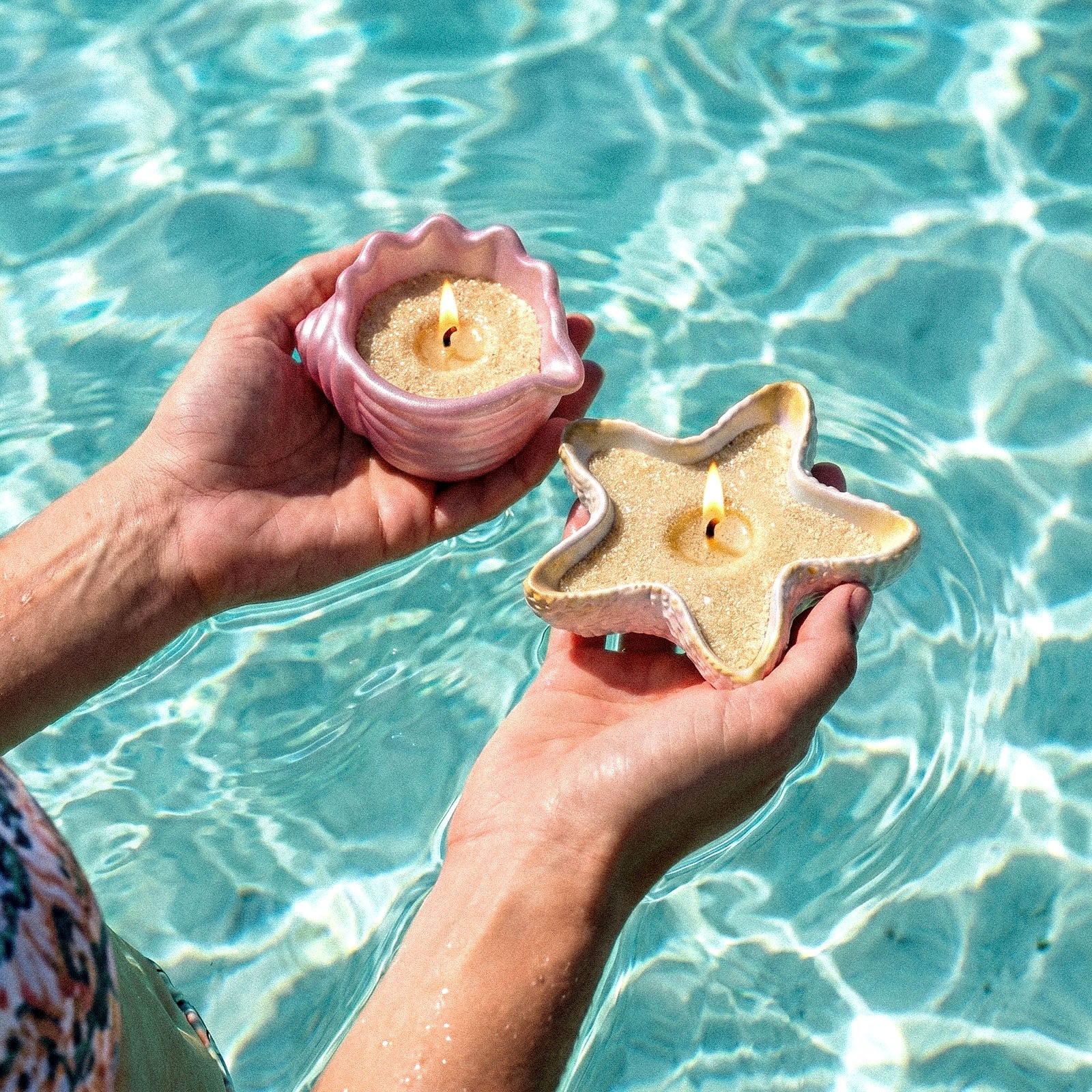 Two Hands Holding Pink & Iridescent White Seashell Sand Wax Candles Over Clear Pool Water - Boowan Nicole