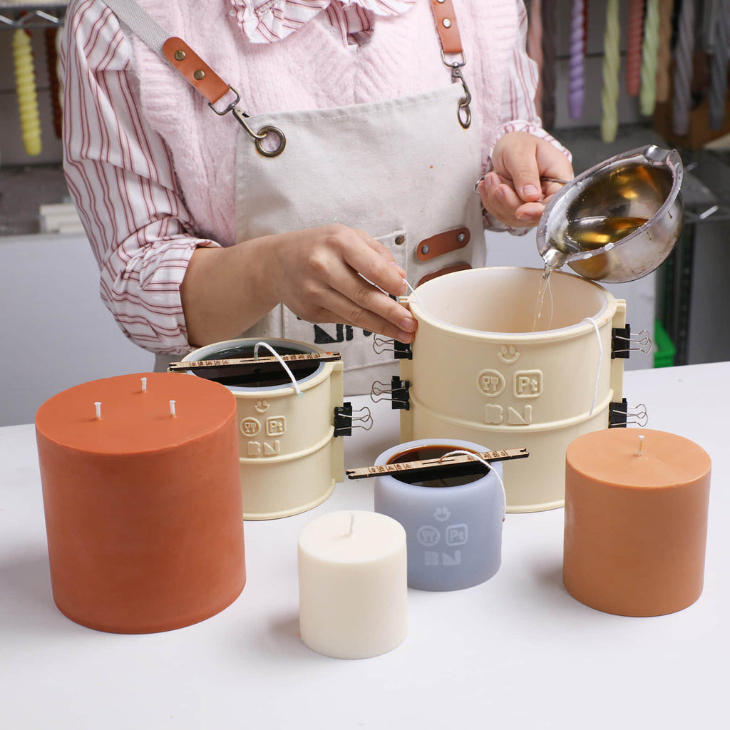 Pouring melted wax into a BN Simple Pillar Candle silicone mold with support shell and wick holders; finished cylinder candles beside - Boowan Nicole