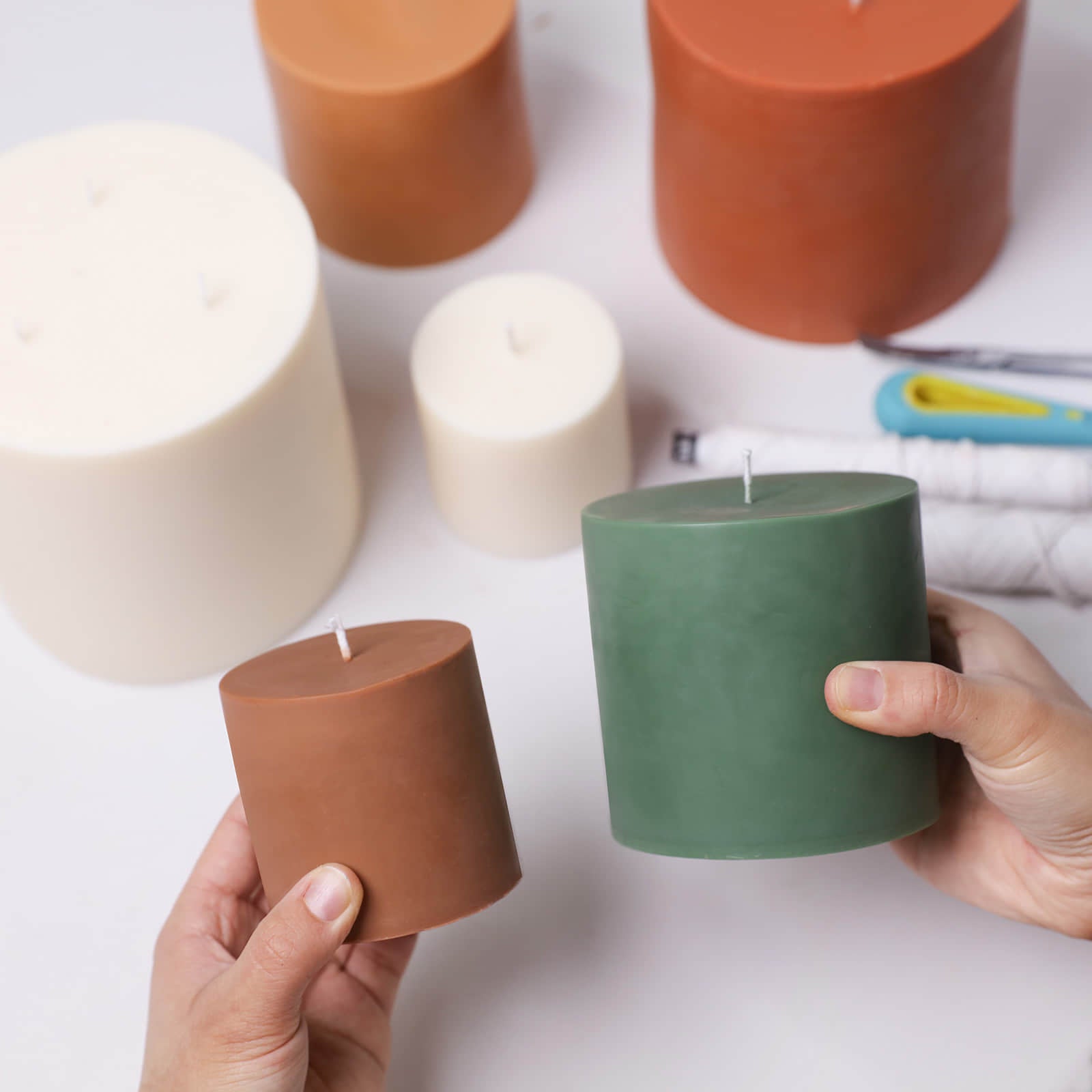Hands holding smooth cylinder pillar candles—matte terracotta and sage green—with larger ivory and terracotta pillars on the worktable - Boowan Nicole