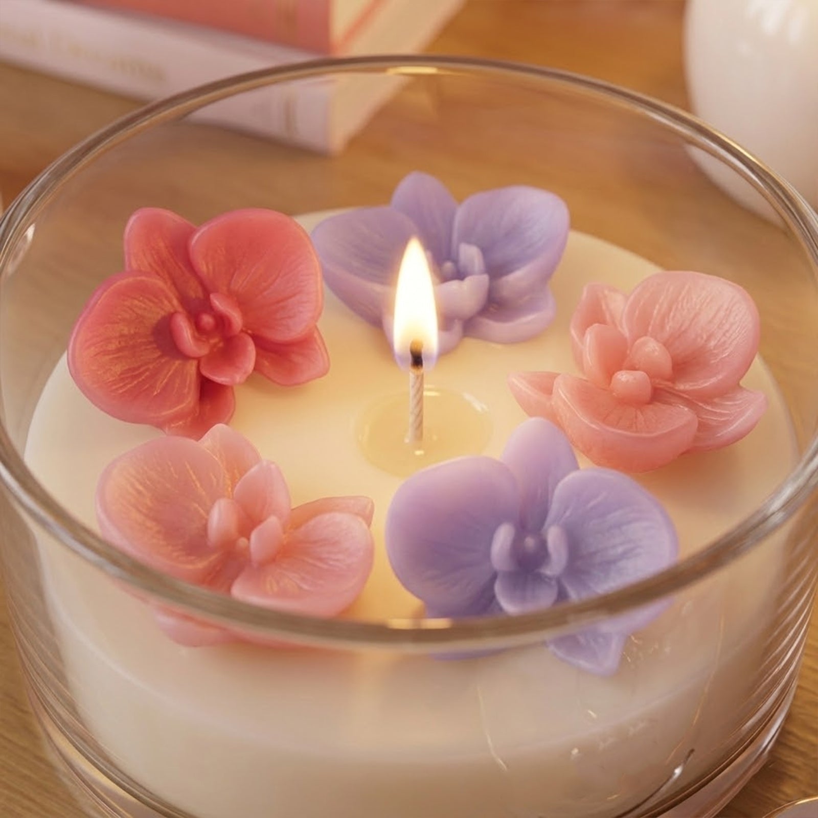 A lit glass candle with pink and purple orchid toppers, casting warm glow and highlighting petal texture. -Boowan Nicole