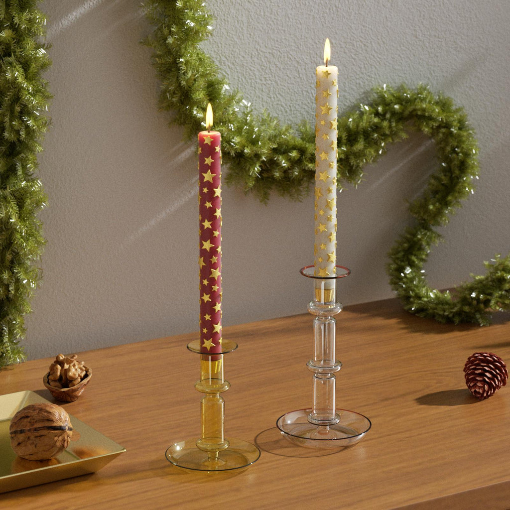 Two festive taper candles with raised golden star patterns, one red and one white, displayed on glass candle holders with Christmas garland in the background - Boowan Nicole