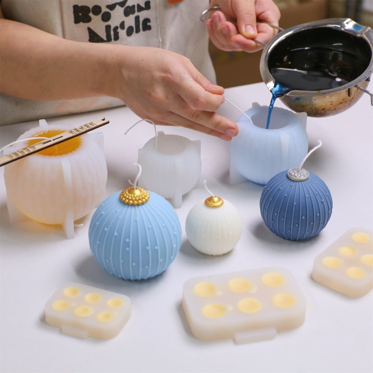 Hand pouring blue wax into Christmas bauble candle silicone mold; finished blue/ivory candles with gold caps and topper-cap molds - Boowan Nicole