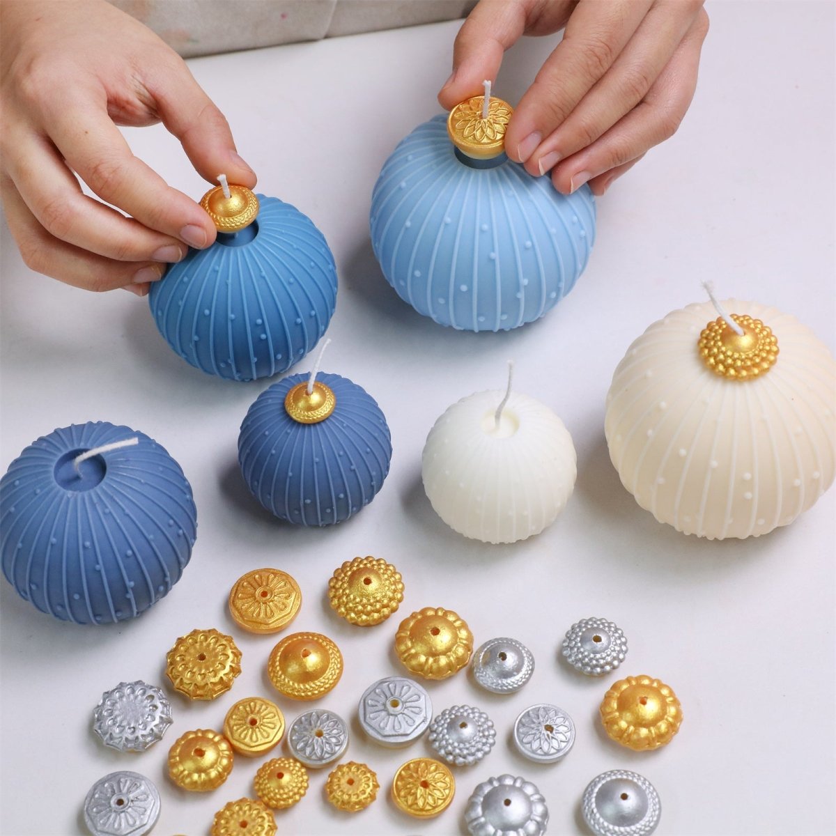 Hands assembling blue and ivory bauble candles with interchangeable gold/silver topper caps displayed - Boowan Nicole