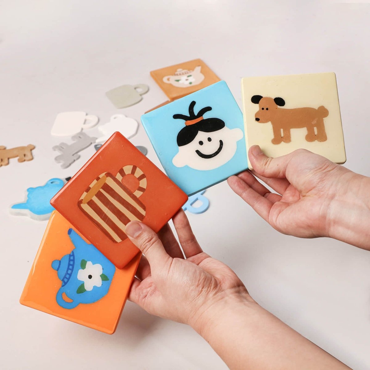 Hands holding colorful square drink coasters with cute mug, face, dog and teapot illustrations - Boowan Nicole