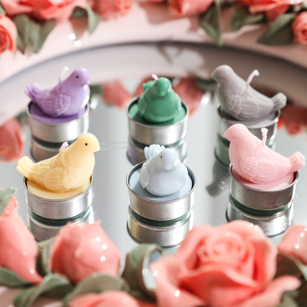 Six dove-shaped tea light candles in different colors, placed on a tray among flowers, designed by Boowan Nicole.
