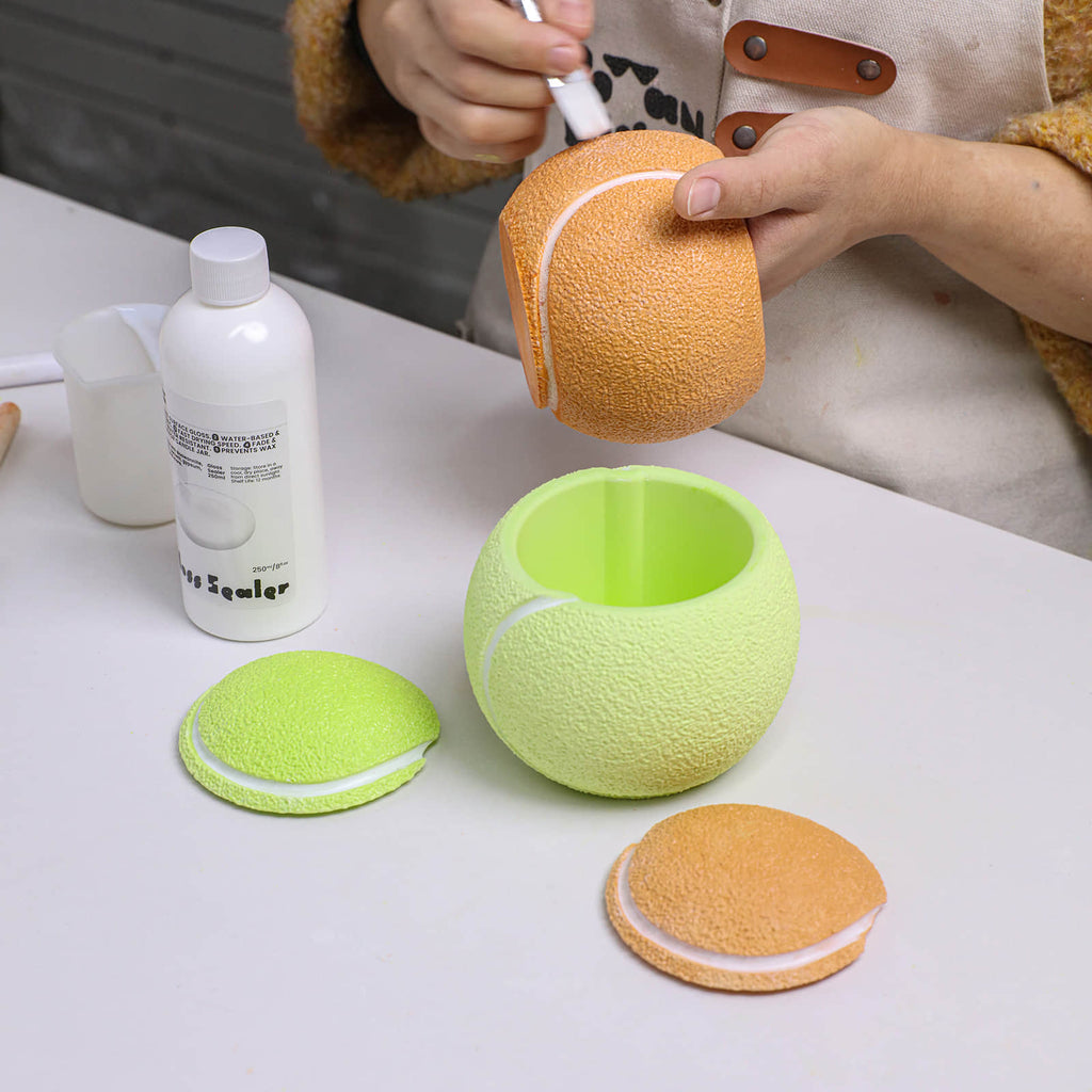 Hand-painting the white seam on an orange tennis-ball candle jar; lime green jar and textured lids on the table, varnish bottle nearby - Boowan Nicole