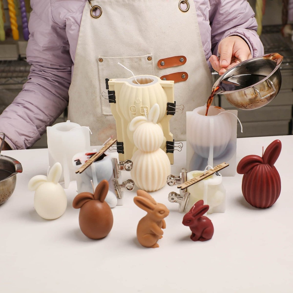 A craftsperson pouring melted wax into silicone molds to create hand-poured ribbed bunny candles, with finished bunny candle designs displayed nearby. - Boowan Nicole