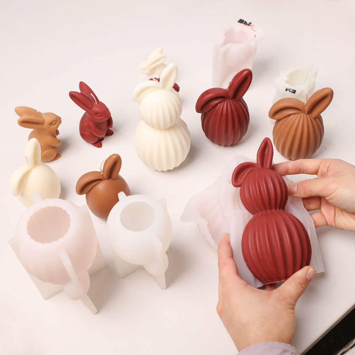 A collection of assorted ribbed bunny candles in cream, burgundy, and brown, with a person removing a finished two-piece burgundy bunny candle from its silicone mold. - Boowan Nicole