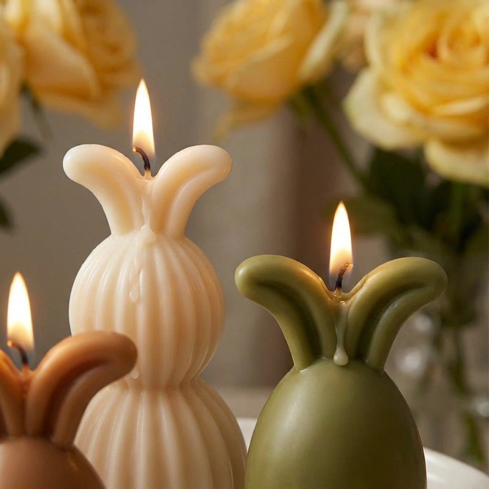 Handcrafted rabbit-shaped pillar candles with warm glowing flames and melting wax, set against a soft-focus background of yellow roses - Boowan Nicole