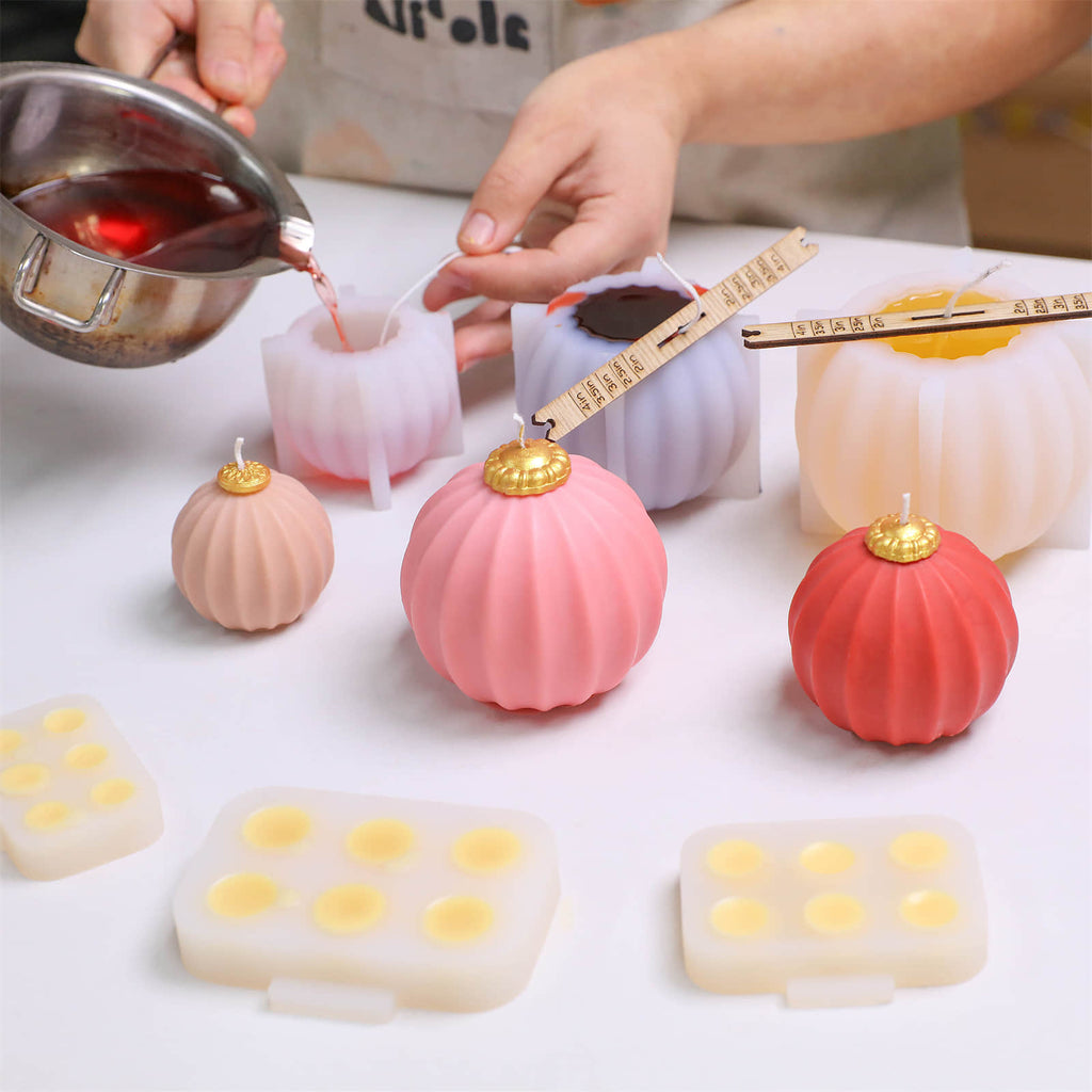 Hand pouring red wax into fluted bauble candle silicone molds; finished pink/red candles with gold caps and topper-cap molds on worktable - Boowan Nicole